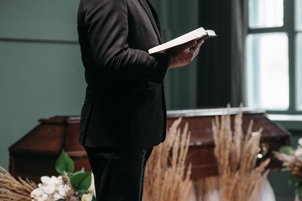 A pastor in a black suit holding a book during a funeral ceremony indoors.