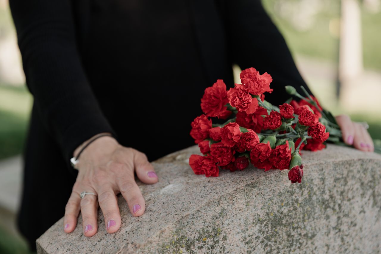 Home A woman dressed in black stands solemnly by a tombstone with red carnations.